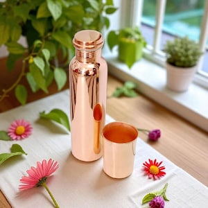 Può includere: Una bottiglia d'acqua e un bicchiere abbinato color rame sono su un panno bianco, che riflette la luce. La bottiglia ha una forma arrotondata con un tappo a vite. Fiori rosa e rossi sono sparsi intorno alla bottiglia e al bicchiere, con piante verdi sullo sfondo.