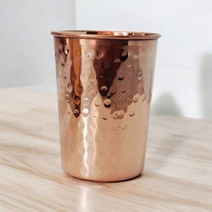 May include: A copper-coloured drinking glass with a hammered texture. The glass has a cylindrical shape with a slightly flared rim. The surface of the glass reflects light. The glass is on a light-coloured wooden surface.