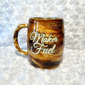 Könnte beinhalten: Eine Kaffeetasse mit einem braunen und beigefarbenen Marmormuster und einem gebogenen Henkel. Auf der Tasse steht in weißer, kursiver Schrift "Maker Fuel". Die Tasse steht auf einer weißen, strukturierten Oberfläche.