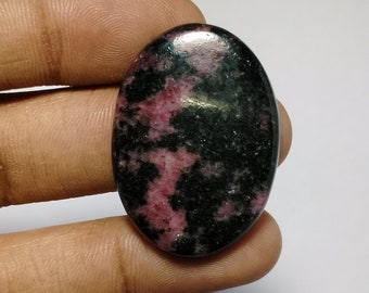 May include: A smooth, oval-shaped cabochon gemstone with a black and pink marbled pattern. The stone is polished and has a glossy finish.