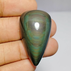 Puede incluir: Una piedra preciosa pulida en forma de lágrima con un patrón arremolinado de tonos verdes, marrones y negros. La piedra tiene un acabado brillante y una superficie lisa y redondeada. La piedra preciosa se sostiene en una mano.