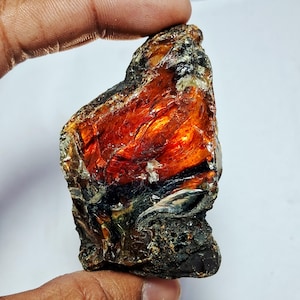 May include: A rough, irregularly shaped amber gemstone with a mix of colors. The stone has a deep reddish-orange hue in the center, transitioning to darker browns and blacks on the edges. The surface appears textured and somewhat translucent.