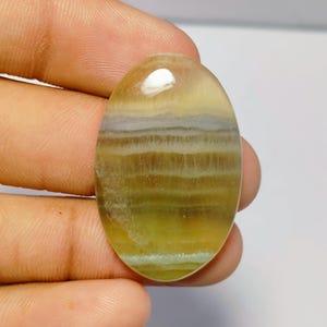 Könnte beinhalten: Ein ovaler Edelstein mit Schichten aus Gelb, Braun und Weiß. Die polierte Oberfläche reflektiert das Licht und zeigt die natürlichen Muster und Farben des Steins. Der Edelstein wird in einer Hand gehalten.