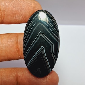 May include: An oval-shaped gemstone with a polished surface. The stone features a dark gray base with intricate white and light gray chevron-like patterns. The gemstone is held between two fingers.