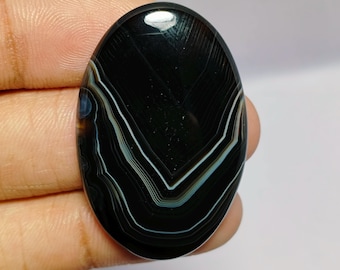 May include: An oval-shaped black agate gemstone with white and light blue banding. The polished stone is held in a hand, showcasing its natural patterns and glossy surface. The gemstone is a striking example of natural stone artistry.