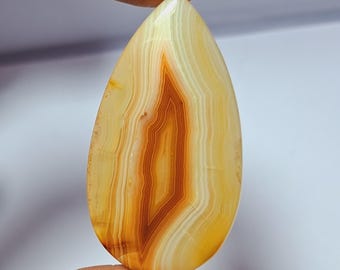 May include: A teardrop-shaped gemstone with a polished surface. The stone features layers of creamy yellow, white, and brown hues, creating a banded pattern. The central area has a rich, reddish-brown color.