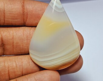 May include: A teardrop-shaped gemstone with a polished surface. The stone features layers of white, cream, and yellow hues, creating a banded pattern. The gemstone is held in a hand, showcasing its size and shape.