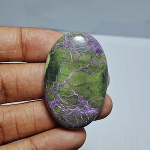 May include: Oval-shaped gemstone with a vibrant green base color, crisscrossed by striking purple veins. The polished stone displays a smooth surface and a natural, organic pattern. The gemstone is held in a hand.