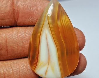 May include: A teardrop-shaped gemstone with a polished surface. The stone features a striking pattern of alternating bands of orange and white, creating a layered effect. The gemstone is held in a hand.