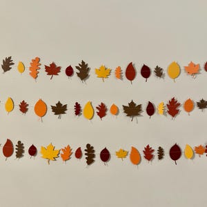 Könnte beinhalten: Drei Girlanden mit Herbstblättern in Rot-, Orange-, Gelb- und Brauntönen. Die Blätter sind in verschiedenen Formen und Größen ausgeschnitten und für eine festliche Herbstdekoration aufgereiht.
