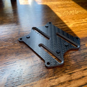 Peut inclure: Une plaque en fibre de carbone noire avec cinq œillets en caoutchouc noir. La plaque comporte une série de fentes et de trous.