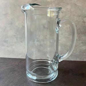 May include: A clear glass pitcher with a handle. The pitcher has a rounded base and a pouring spout. The handle is curved and attached to the side of the pitcher. The pitcher is empty and sits on a dark surface.