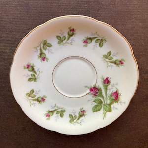 Könnte beinhalten: Eine weiße Untertasse mit goldfarbenem Rand und einem Blumendesign mit rosa Rosen und grünen Blättern.