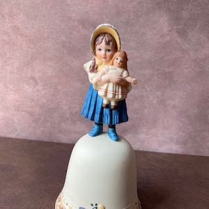 Könnte beinhalten: Eine Porzellan-Glocke mit einer Figur eines Mädchens, das eine Puppe hält. Das Mädchen trägt ein blaues Kleid und einen weißen Hut. Die Glocke ist mit einem blauen Band und Blumenmustern verziert.