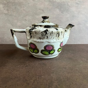 Peut inclure: Une théière en céramique blanche avec un glaçage moucheté brun et des motifs floraux roses et verts. La théière a un couvercle et un bec.