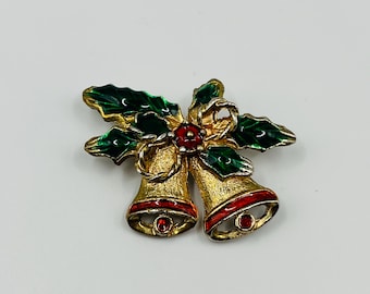 May include: A gold-toned Christmas bell brooch with red accents and green holly leaves. The brooch features two bells with red rims and a red gem at the base of each. The holly leaves are green with gold detailing.