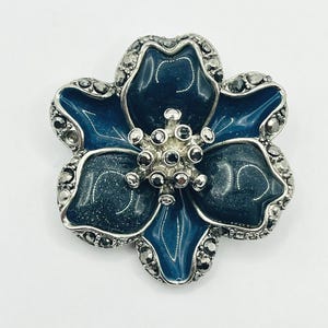 May include: A flower-shaped brooch with a silver-tone metal frame. The brooch features a dark blue enamel center and petals, with a silver-tone center and accents. The brooch is a jewelry accessory.