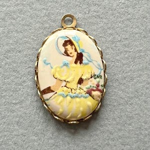 May include: Vintage oval pendant with a gold-tone frame. The pendant features a painted image of a woman in a yellow dress and a blue bonnet, holding a basket of flowers. The background is cream-colored.