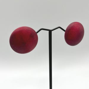 May include: A pair of round, red stud earrings displayed on a black jewelry stand. The earrings have a smooth, matte finish and a deep red color. The stand is simple and elegant, with a vertical post and two horizontal arms.