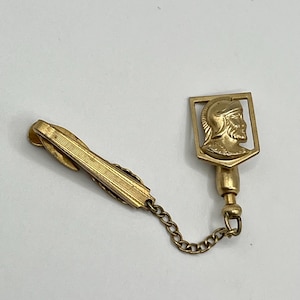 以下が含まれることがあります： 金色のネクタイクリップ。ヘルメットをかぶった男性の横顔が描かれた装飾的なシールドが付いています。クリップはチェーンで装飾要素に接続されています。