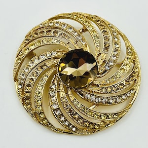 May include: A circular gold-tone brooch featuring a large, faceted, dark brown central stone surrounded by radiating, curved metal arms. These arms are embellished with rows of small, clear and light yellow crystals in a swirling, sunburst pattern.
