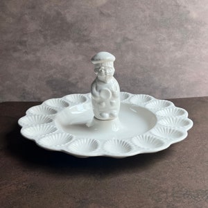 May include: White ceramic egg dish with a sculpted figure of a chef holding a spoon in the center. The dish has 12 individual compartments shaped like seashells.
