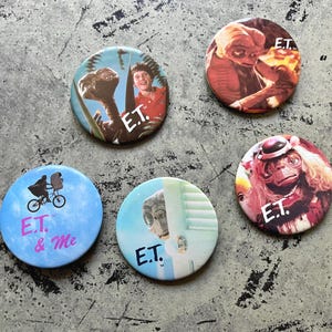 Könnte beinhalten: Eine Sammlung von fünf runden Ansteckbuttons mit Bildern aus dem Film E.T. Der Außerirdische. Die Buttons zeigen verschiedene Szenen und Charaktere, darunter E.T. und den Jungen, sowie den Text "E.T. & Me".