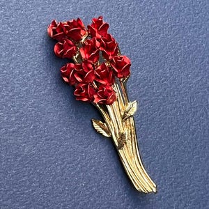 May include: A gold-tone metal brooch shaped like a bouquet of red roses. The piece features multiple deep red enameled rose blossoms clustered at the top, with textured gold stems and small leaves extending downward to form the base of the bouquet.