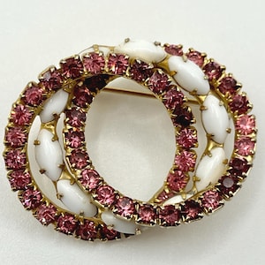 May include: A vintage brooch featuring two interlocking gold-toned circles. Each circle is adorned with small, pink rhinestones and white, oval-shaped accents. The brooch has a pin closure on the back.