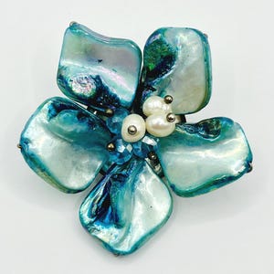 May include: A flower-shaped brooch with iridescent blue and green petals. The center features three white pearls and blue crystal accents. The brooch is made of a shell-like material and has a silver-tone metal backing.