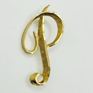 May include: A gold-toned metal brooch in the shape of the uppercase letter P. The letter features a cursive, flowing script design with a textured, brushed finish across its entire surface, creating a reflective and dimensional appearance.
