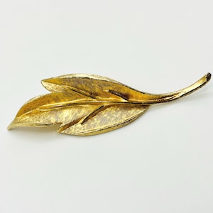 May include: A gold-toned leaf-shaped brooch with textured detailing. The brooch features a long, curved stem and three detailed leaves. The jewelry piece is set against a white background.