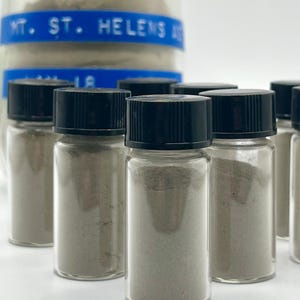 May include: Small glass vials filled with gray powder, each sealed with a black cap. A larger jar with a blue label reading "MT. ST. HELENS ASH" is visible in the background. The vials are arranged in a group, showcasing the product.