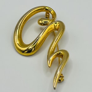 May include: A gold-toned brooch in the shape of the letter 'G'. The brooch is made of two pieces of metal, one gold and one silver, that are joined together to form the letter. The brooch is a simple and elegant design.