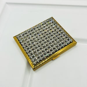 May include: A rectangular compact mirror case featuring a gold-toned metal frame and a top surface densely covered in clear, faceted rhinestones arranged in a grid pattern. The case has a hinged closure mechanism visible on the side edge.