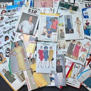 May include: A large collection of vintage sewing pattern envelopes from brands including Simplicity, Vogue, and Butterick. The paper envelopes feature various illustrations of clothing designs, model photographs, brand logos, and printed text labels.