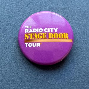 May include: A round, purple button with the text "THE RADIO CITY STAGE DOOR TOUR" in yellow and white. The button is set against a blue textured background. The button is a souvenir or promotional item.