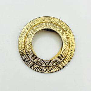 May include: A gold-toned brooch with a textured, concentric circle design. The brooch features a repeating pattern of small, diamond-shaped indentations across its surface. The circular shape is a classic design element.