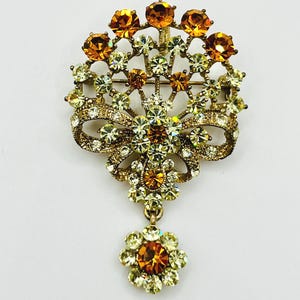 May include: A vintage brooch with a floral design. The brooch features a bow and a dangling flower, all adorned with amber and clear rhinestones. The metal has a gold tone. The brooch is a classic piece of jewelry.