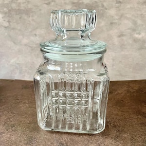 Op de afbeelding: Een heldere glazen pot met een deksel. De pot heeft een vierkante vorm met verticale en vierkante textuurpatronen. Het deksel heeft een decoratief, geribbeld ontwerp. De pot staat op een bruin oppervlak.