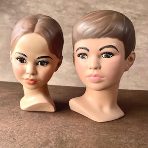 May include: Two vintage mannequin heads with painted features. One has a side part and the other has a short, boyish haircut. Both have painted eyes, eyebrows, and lips. The heads are a flesh tone colour.