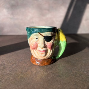 May include: A ceramic mug shaped like a pirate's head with a green eye patch and a parrot perched on the handle. The mug is painted in shades of brown, green, and yellow.