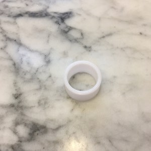 May include: A white, cylindrical object with a hollow center, resting on a marble surface. The object appears to be made of a solid, opaque material. The marble surface has gray and black veining.