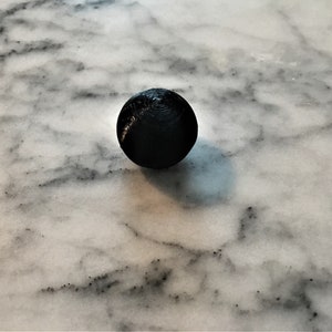 May include: A black sphere with a textured surface sits on a white marble background.