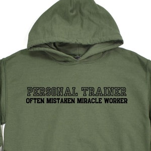 Könnte beinhalten: Ein grüner Kapuzenpullover mit dem Aufdruck "PERSONAL TRAINER OFTEN MISTAKEN MIRACLE WORKER" auf der Vorderseite.