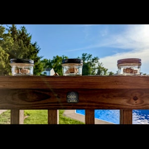 May include: Three small glass jars with black lids filled with wooden candle melts. The jars are sitting on a wooden railing with a blue swimming pool in the background. The jars have a label that says "Calvin Candle".