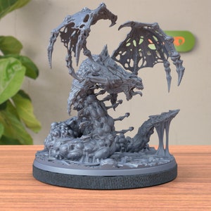 May include: A gray, detailed sculpture of a monstrous creature with large wings and a serpentine body. The sculpture has a textured surface and is mounted on a round, black base. The creature has an open mouth and sharp teeth.