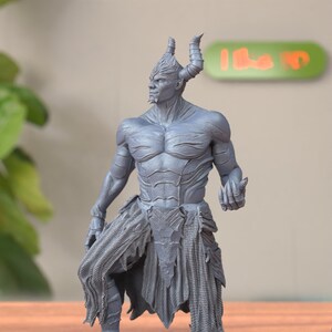 May include: A gray 3D printed figurine of a demon with horns, a muscular build, and a tattered skirt. The demon is standing with one leg bent and the other straight, and its right hand is raised.
