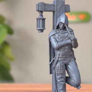 May include: A gray 3D printed miniature of a hooded figure leaning against a wooden post with a lantern hanging from it. The figure is holding a dagger in one hand and has a belt with pouches on their hip.