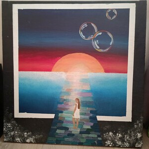 Puede incluir: Pintura original que representa a una mujer con un vestido blanco de pie en un camino de piedra que conduce a una puesta de sol sobre el agua. Burbujas flotan en el cielo. La pintura está enmarcada con un borde blanco y un marco exterior negro.